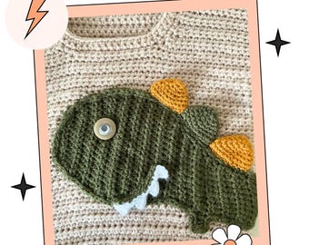 May include: Crocheted beige sweater with a green dinosaur applique. The dinosaur has yellow spikes, white teeth, and a button eye. The sweater is framed with a pink border and decorated with a lightning bolt, stars, and a flower.