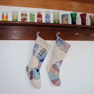 May include: Two fabric Christmas stockings hanging from a wooden peg rack. Each stocking features a cream-colored base with a fan-shaped patchwork design made of various blue, white, and patterned fabric scraps stitched onto the front surface.