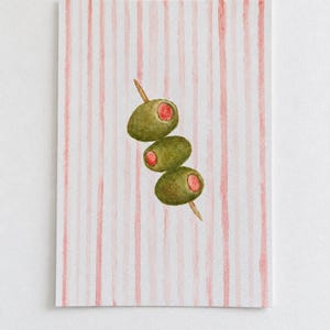 May include: A watercolour painting on paper featuring three green olives with red pimento centres skewered on a wooden toothpick. The background consists of vertical, hand-painted light pink stripes on a white surface, creating a simple composition.