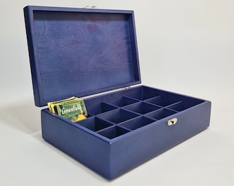 Könnte beinhalten: Eine blaue Holzteekiste mit einem Scharnierdeckel und einem silbernen Verschluss. Die Kiste hat neun Fächer zum Aufbewahren von Teebeuteln. In einem der Fächer ist eine Schachtel "Greenfield Black Tea" Teebeutel sichtbar.