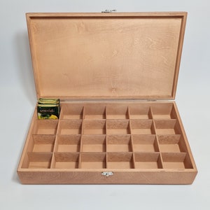 May include: A wooden box with a hinged lid and a latch. The box has 24 compartments for storing tea bags. The box is open and there are tea bags in one of the compartments. The tea bag packaging says "Greenfield" and "Lemon".