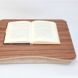 Può includere: Un tavolino da letto in legno marrone con un cuscino in tessuto beige. Il tavolino ha un bordo arrotondato ed è progettato per essere utilizzato su un letto o un divano. Un libro aperto è posto sul tavolino.
