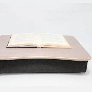 Puede incluir: Un escritorio de madera gris y marrón para ordenador portátil con un cojín de tela negro. El escritorio está apoyado en el cojín, creando una superficie cómoda para trabajar o leer. Un libro está colocado en el escritorio.