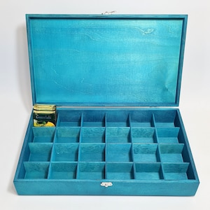 Puede incluir: Caja de madera de color turquesa con tapa con bisagras y 24 compartimentos. La caja está abierta y se puede ver una caja de bolsitas de té en uno de los compartimentos. La caja de bolsitas de té dice "Greenfield Lemon".