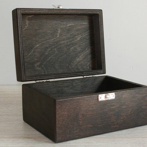 Puede incluir: Una caja de madera marrón oscuro con una tapa con bisagras y un cierre plateado. La caja está abierta y vacía.