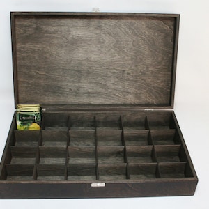 May include: A dark brown wooden tea box with a hinged lid and a compartment for storing tea bags. The box has 24 individual compartments for storing tea bags. The box is open and a tea bag is visible in one of the compartments. The tea bag is labeled "Greenfield Lemon".