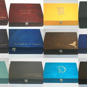 May include: Twelve wooden boxes with various colors and engraved text. The boxes have a hinged lid and a metal clasp. The text on the boxes includes quotes, names, and a logo.