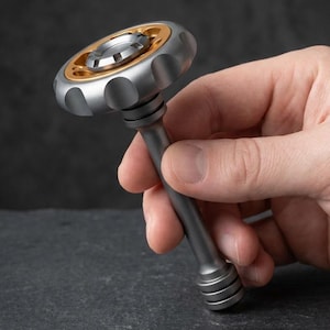 May include: A metal fidget spinner tool featuring a matte silver finish with a gold-toned circular accent plate. The device has a long, cylindrical handle and a top section with scalloped edges, designed for rotation and tactile sensory engagement.