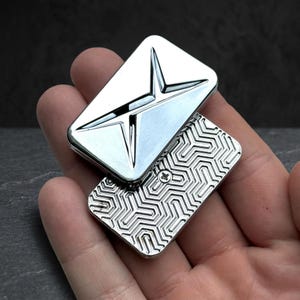 May include: A rectangular silver-coloured metal object with a smooth, polished top surface featuring a recessed star-like design. The bottom section has a textured, repeating geometric pattern and a single visible screw head in the centre.