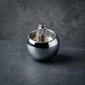 Puede incluir: Una peonza de metal de color plateado pulido con un cuerpo esférico y un vástago vertical central. El vástago presenta un patrón de agarre moleteado y texturizado en la parte superior, mientras que el cuerpo principal tiene un acabado metálico liso y reflectante.