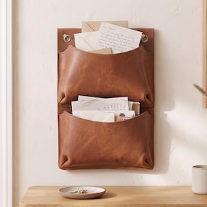 May include: A wall-mounted brown leather organiser featuring two stacked pockets for storing mail and documents. The organiser is secured with two silver-toned metal grommets at the top and contains several white envelopes and papers.