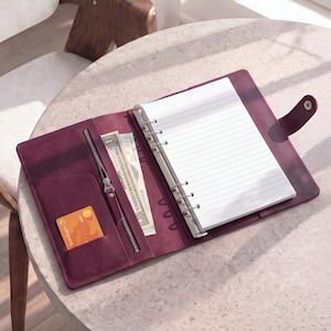 Peut inclure: Un agenda en cuir violet foncé doté d'un mécanisme de reliure métallique à six anneaux contenant du papier blanc ligné. L'intérieur comprend une poche zippée avec un billet d'un dollar et une fente pour une carte bancaire orange.