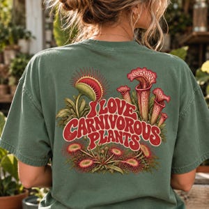Puede incluir: Una camiseta verde salvia con un gráfico en la espalda que incluye el texto I LOVE CARNIVOROUS PLANTS en letras rojas. El diseño presenta ilustraciones de una venus atrapamoscas, plantas jarro y droseras en tonos rojos, verdes y amarillos.