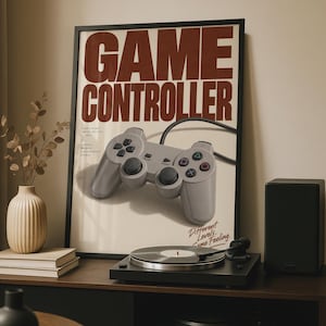May include: A framed poster featuring the text GAME CONTROLLER in large dark red letters above a light grey wired video game controller. The poster includes smaller text details and the phrase Different Levels, Game Feeling in red script.