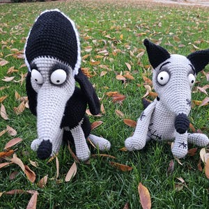Puede incluir: Dos animales de peluche tejidos a mano. Uno es un perro negro y gris claro con grandes ojos blancos y un hocico largo. El otro es un perro gris claro con orejas negras, nariz negra, grandes ojos blancos y detalles cosidos en el cuerpo.