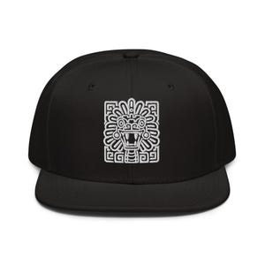 May include: A black snapback cap featuring a white embroidered graphic of a stylized Mesoamerican deity face with geometric patterns and an open mouth with visible teeth centered on the front panel of the hat.