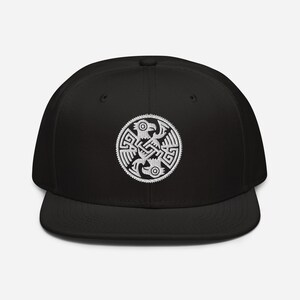 May include: A black baseball cap featuring a white embroidered circular emblem on the front. The emblem displays a detailed, symmetrical design of two stylized birds facing each other, integrated with intricate geometric knotwork patterns.