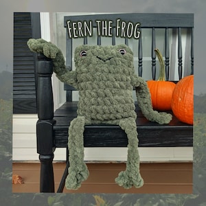 May include: A plush olive green frog toy with a textured, chunky knit appearance, featuring small dark eyes and a stitched mouth. The text "FERN THE FROG" is displayed above the toy, which is positioned next to two orange pumpkins.