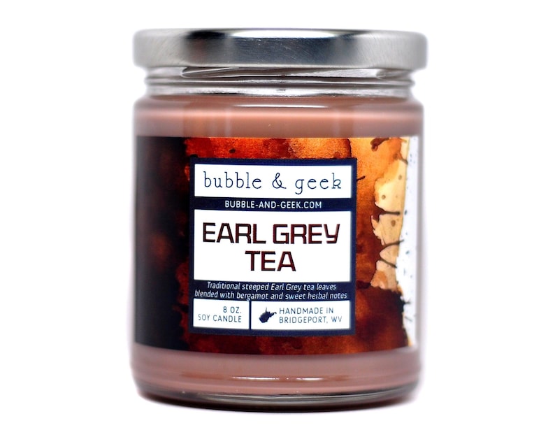 Op de afbeelding: Een glazen potkaars met een bruin etiket waarop "bubble & geek" en "EARL GREY TEA" staat. Het etiket vermeldt ook "Traditional steeped Earl Grey tea leaves blended with bergamot and sweet herbal notes". De kaars is 8 oz en is een sojawas kaars. Hij is handgemaakt in Bridgeport, WV.