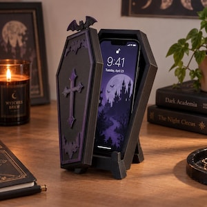 May include: A black coffin-shaped smartphone stand featuring a purple cross and bat accents on the lid. The stand holds a smartphone displaying a screen with the time 9:41, the date Tuesday, April 23, and a graphic of a castle under a full moon.