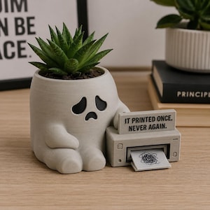 Puede incluir: Una maceta de color gris claro con forma de fantasma y una suculenta verde, que presenta una cara triste y un modelo de impresora pequeño. La impresora tiene el texto IT PRINTED ONCE, NEVER AGAIN. y una hoja con un garabato negro.