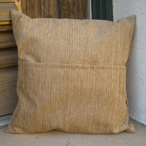 May include: A square, tan corduroy pillow with a vertical ribbed texture. The pillow has a sewn seam across the middle and a flap on the back. The pillow is resting on a concrete surface.