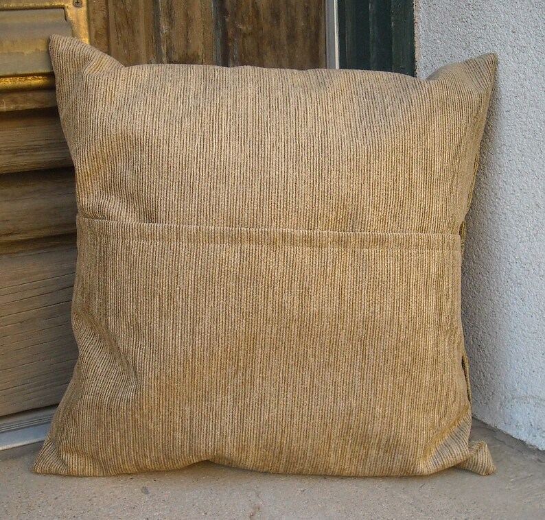 May include: A square, tan corduroy pillow with a vertical ribbed texture. The pillow has a sewn seam across the middle and a flap on the back. The pillow is resting on a concrete surface.