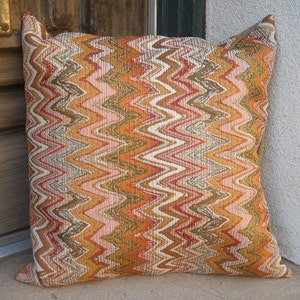 May include: Square decorative pillow with a zig-zag pattern in shades of orange, brown, pink, and white. The pillow has a textured appearance and is leaning against a wall.