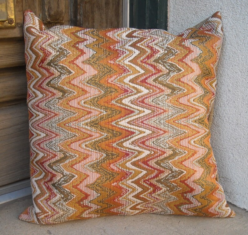 May include: Square decorative pillow with a zig-zag pattern in shades of orange, brown, pink, and white. The pillow has a textured appearance and is leaning against a wall.