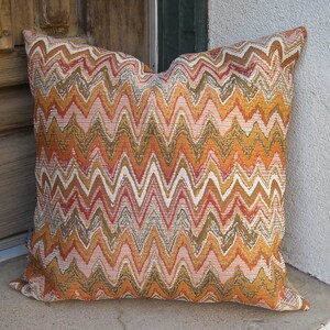 May include: Square decorative pillow with a vibrant zigzag pattern in shades of orange, white, red, and brown. The pillow is made of a textured fabric and appears to be filled with a soft material, suitable for home decor.