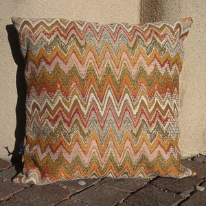 May include: Square decorative pillow with a woven chevron pattern in shades of orange, gold, pink, and white. The pillow is placed on a brick surface and leans against a beige wall.