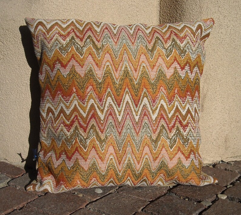 May include: Square decorative pillow with a woven chevron pattern in shades of orange, gold, pink, and white. The pillow is placed on a brick surface and leans against a beige wall.