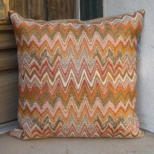 May include: Square decorative pillow with a zig-zag pattern in shades of orange, brown, pink, and white. The pillow has a textured appearance and a neutral-colored border. The pillow is resting on a concrete surface.