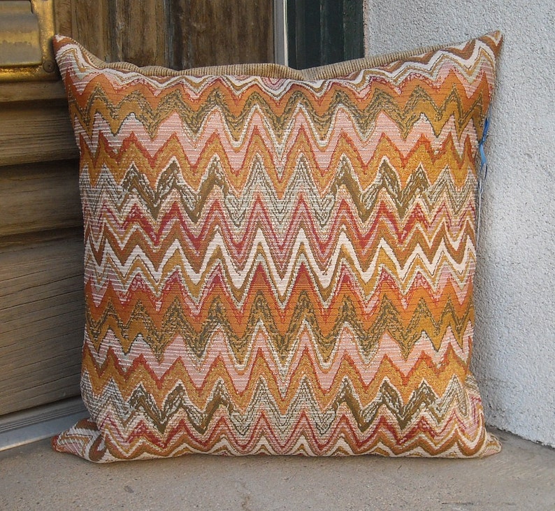May include: Square decorative pillow with a zig-zag pattern in shades of orange, brown, pink, and white. The pillow has a textured appearance and a neutral-colored border. The pillow is resting on a concrete surface.