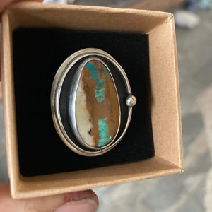 Op de afbeelding: Zilveren ring met een ovale steen in bruin, wit en turquoise tinten. De ring heeft een dubbele zilveren band en een klein zilveren kogeldetail. Gepresenteerd in een kleine, open kartonnen doos met een zwart interieur.