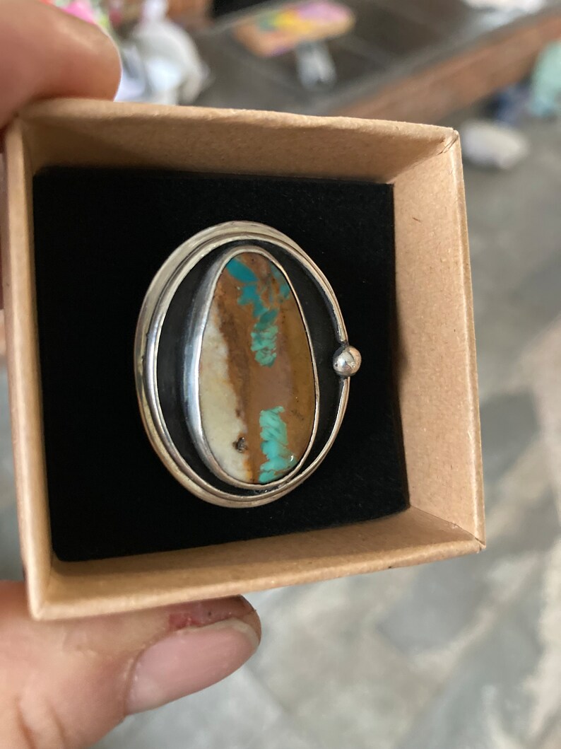 Op de afbeelding: Zilveren ring met een ovale steen in bruin, wit en turquoise tinten. De ring heeft een dubbele zilveren band en een klein zilveren kogeldetail. Gepresenteerd in een kleine, open kartonnen doos met een zwart interieur.