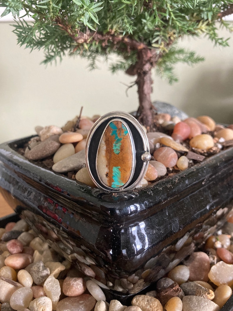 Op de afbeelding: Ovale zilveren ring met een grote, natuurlijke steen in bruin-, beige- en turkooistinten. De steen is gezet in een zwarte en zilveren bezel, wat een opvallend contrast cre&euml;ert. De ring wordt gepresenteerd op kleine stenen.