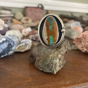 Op de afbeelding: Een zilveren ring met een ovale steen. De steen heeft bruine, turquoise en cr&egrave;mekleurige tinten. De ring ligt op een donkergrijze rots, met een houten oppervlak op de achtergrond.