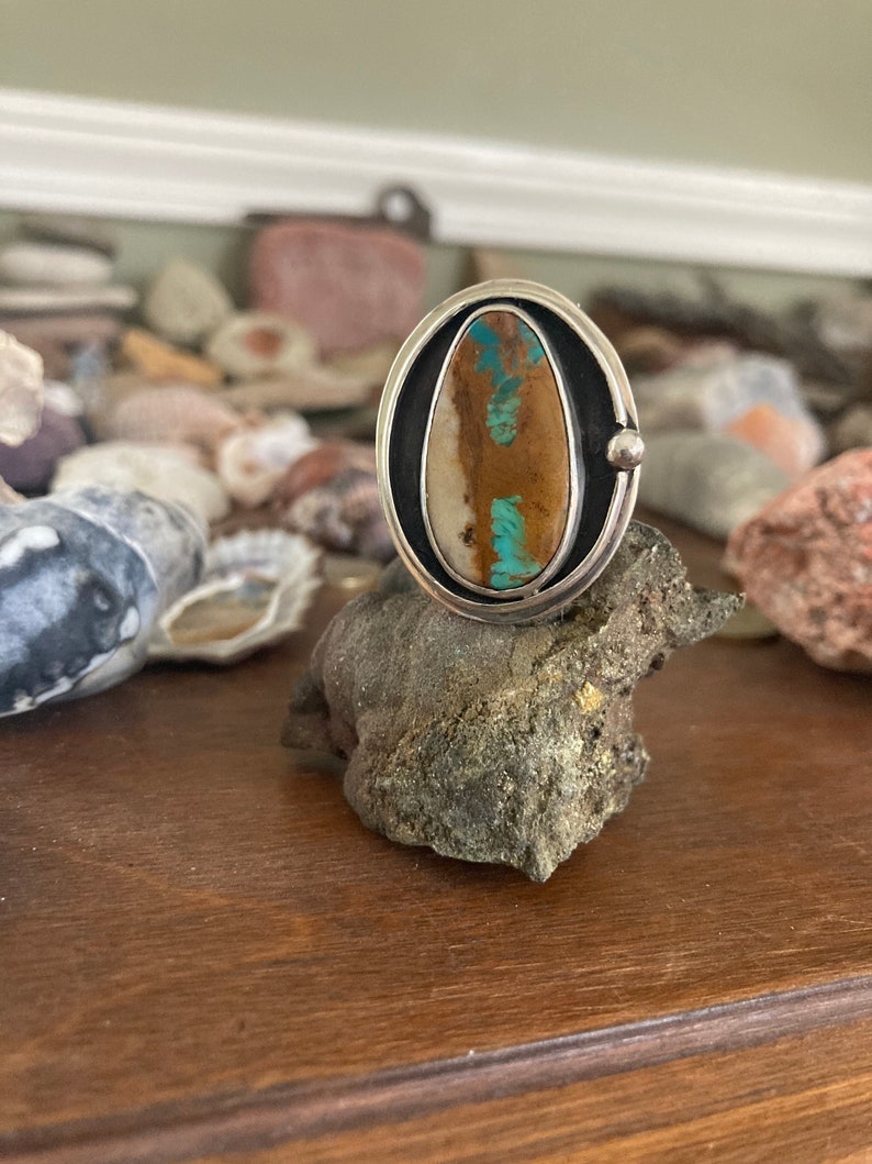 Op de afbeelding: Een zilveren ring met een ovale steen. De steen heeft bruine, turquoise en cr&egrave;mekleurige tinten. De ring ligt op een donkergrijze rots, met een houten oppervlak op de achtergrond.
