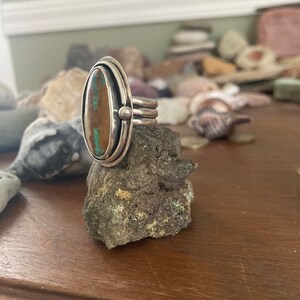 Op de afbeelding: Een zilveren ring met een ovaalvormige turkoois steen. De ring heeft een dubbel bandontwerp met een kleine zilveren kraal aan de zijkant. De steen is een mix van bruin en blauwgroen. De ring wordt gepresenteerd op een kleine rots.