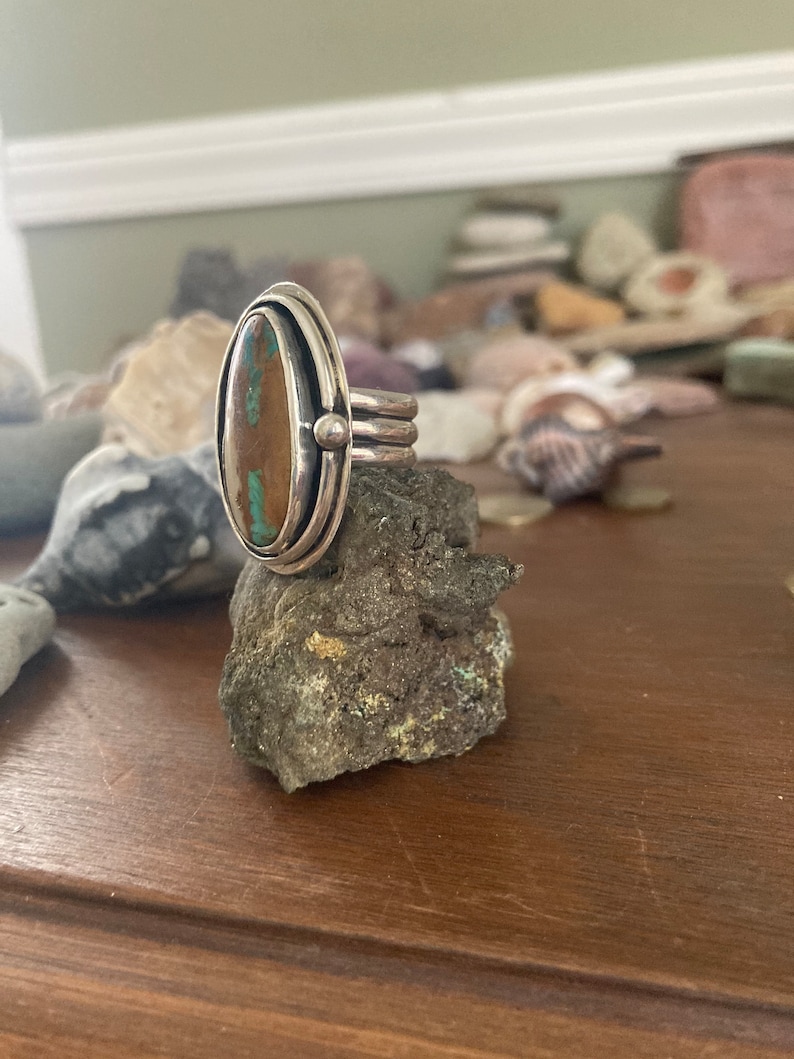 Op de afbeelding: Een zilveren ring met een ovaalvormige turkoois steen. De ring heeft een dubbel bandontwerp met een kleine zilveren kraal aan de zijkant. De steen is een mix van bruin en blauwgroen. De ring wordt gepresenteerd op een kleine rots.