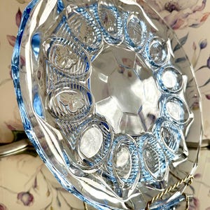 May include: A round, light blue glass plate featuring a central octagonal panel surrounded by a ring of twelve oval indentations. Each oval indentation contains a raised floral motif, and the outer edge of the plate has a scalloped design.