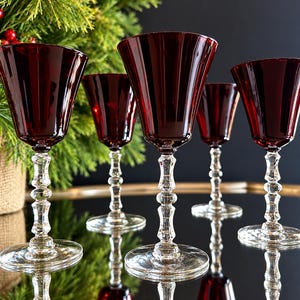 May include: A set of five wine glasses featuring deep red, translucent bowls with a flared rim. Each glass has a clear, multi-faceted, decorative stem and a circular base. The glasses are arranged in a group, reflecting on a dark surface.