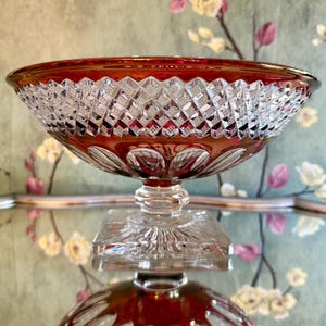 May include: A red and clear cut glass pedestal bowl featuring a diamond-patterned rim and a scalloped base design. The bowl sits on a square clear glass foot, showcasing intricate geometric detailing and a vibrant ruby-colored upper section.