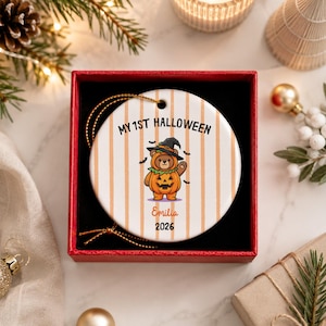 May include: A round white ceramic ornament with vertical orange stripes, featuring a cartoon bear in a pumpkin costume and witch hat. The text reads MY 1ST HALLOWEEN, Emilia, and 2026. It includes a gold hanging string and sits in a red gift box.