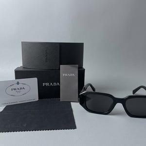 Può includere: Occhiali da sole rettangolari neri con montatura spessa, accompagnati da una custodia nera marchiata, un piccolo cartoncino informativo grigio, un certificato di autenticità bianco e un panno in microfibra nero con bordo seghettato.