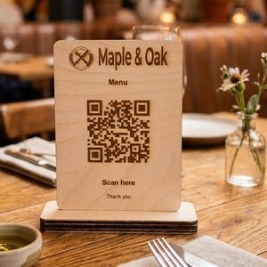 May include: A rectangular light-coloured wooden menu sign with a dark brown engraved QR code, the text Maple & Oak, Menu, Scan here, and Thank you. The sign is mounted on a matching wooden base and stands upright on a wooden table surface.