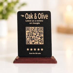 Puede incluir: Un letrero rectangular de madera negra sobre una base de color rojo oscuro. El letrero presenta el texto Oak & Olive, Leave us a review on Google, un código QR central, cinco estrellas doradas y el texto Scan the QR code en letras doradas.