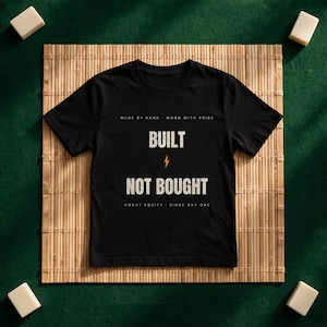 May include: A black short-sleeve t-shirt featuring the text "MADE BY HAND - WORN WITH PRIDE" at the top, "BUILT" in large letters, a small orange lightning bolt icon, "NOT BOUGHT" in large letters, and "SWEAT EQUITY - SINCE DAY ONE" below.