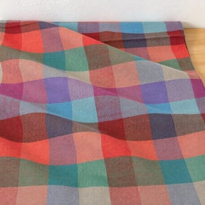 Op de afbeelding: Een textielstof met een meerkleurig ruitpatroon met herhalende vierkanten in tinten rood, groenblauw, lichtblauw, paars en beige. Het materiaal is gedrapeerd om een gestructureerd oppervlak met een consistent rasterontwerp door de stof te tonen.